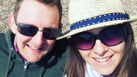 Tom Youngs is on the left and sitting next to Michelle Youngs, who is on the right. He has short brown hair and is wearing black sunglasses and a green jacket. She has long brown hair and is wearing a beige hat with a blue pattern and sunglasses. They are sitting on a beach. 