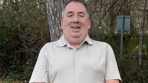 A man wearing a beige shirt is standing in a garden. He has tattoos all over his arms and is wearing a watch.