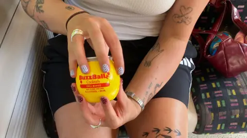 Someone holding a yellow BuzzBallz cocktail container, it has red writing on and says Chili Mango. The person holding it has inverted polka dot nails, on one hand white and red, on the other red and white. They are sat on a train.