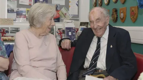 An elderly man is sitting on a chair wearing a suit and tie. He is smiling and holding a walking stick and piece of paper in his hand. Next to him is a woman in a pink jumper smiling at him.