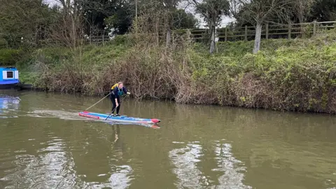 A stand-up paddleboarder goes along a canal, a green bank in the background