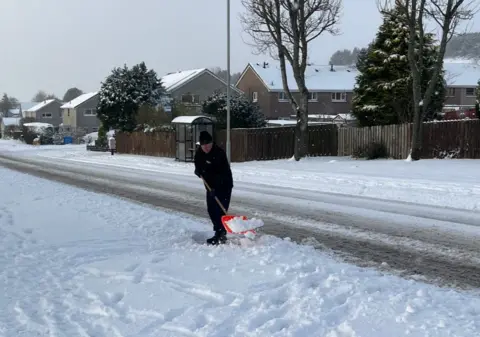 A man dressed in warm, black clothing clearing snow from a pavement in a suburban residential area