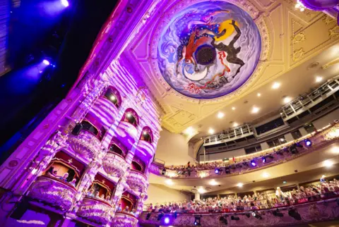 Anneleen Lindsay The restored auditorium of Edinburgh's King's Theatre is illuminated in purple and blue light as audience members fill the stalls, balconies and upper circle. The theatre's ornate decorative interior and the dome painting All the World's a Stage by John Byrne are visible above the auditorium.