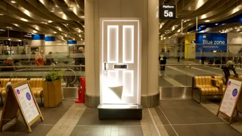 A front door on the concourse at a railway station, next to platform 5a. The white door has a light shining from underneath