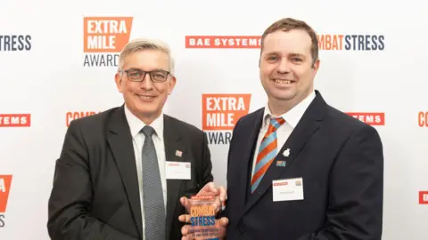 Combat Stress Two men smiling while looking straight at the camera. They are stood in front of an advertising banner. They are both wearing suits and holding an award.