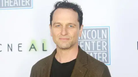 Matthew Rhys wearing a black top and brown blazer. He stands in front of a white background with several logos. 