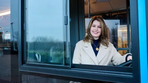 North East Combined Authority Kim McGuinness sitting behind the steering wheel of a bus. She is wearing a white coat and a blue top. She has one hand on the steering wheel and is looking out of the window and smiling at the camera.