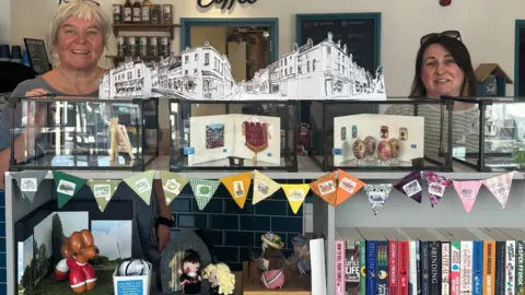 Two women stand behind shelves - on top is a row of clear shoeboxes with the displays in them. Bunting surrounds with a shelf underneath having knitted figures.
