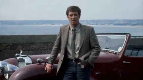 John Nettles is standing in front of a burgundy Triumph Roadster car. He’s resting a hand on the car that is parked in front of a sea wall in Jersey with a view of a bay behind it. He's wearing dark blue jeans with a belt and a shirt and tie with a tweedy grey jacket.