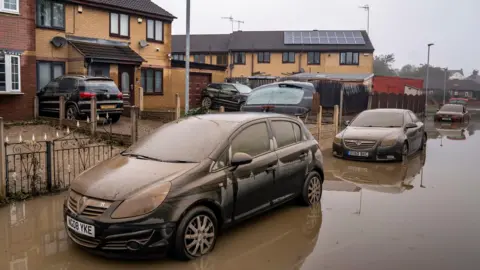 Danny Lawson/PA Wire Cars covered in silt stand in floodwater on a street
