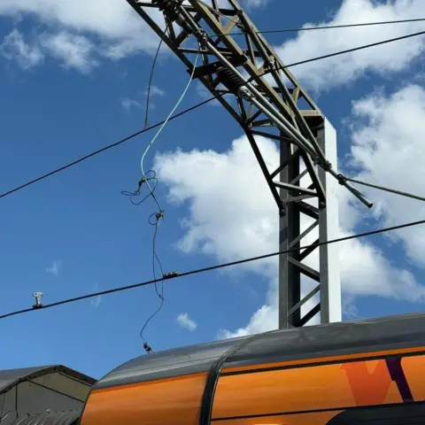 Network Rail Loose cables hanging near a steel beam with the roof of a train showing below them. The sky is blue overhead with some white clouds.