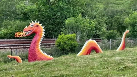 Malcolm Curley The image shows a sculpture of a red, orange and white dragon disappearing into the grass. It looks like it is blowing fire from it's mouth. There are railways tracks behind the dragon and trees in the background.
