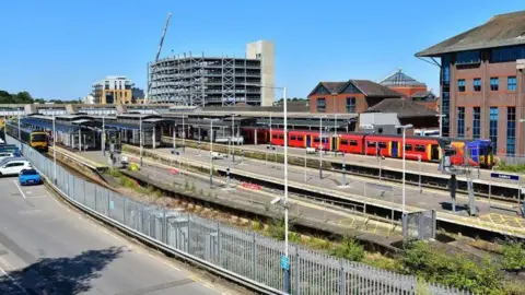 Network Rail Guildford Railway Station. There is clear blue skies, a red train stopped at the station and a car park.