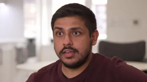 A man with short brown hair, a short brown moustache and beard wearing a maroon T-shirt. He is sitting in a light home.