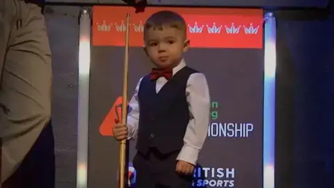 Luke stands on a stage wearing a black waistcoat over a white shirt. He wears a red bowtie and holds a small snooker cue 