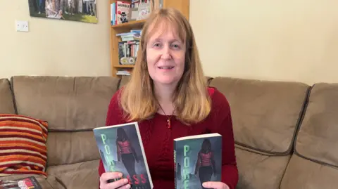 George King/BBC A woman with shoulder-length hair sitting on a brown sofa while holding two copies of her book Powerless. She is looking into the camera