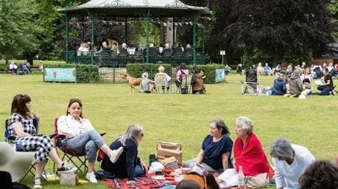 Stu Vincent Photography/Northampton Town Council The park with a bandstand in the background, on which musicians are playing. There are women gathered around a blanket in the foreground - two are sitting on folding chairs while five are sitting on the blanket or grass. There is a low hedge around the bandstand and trees behind it.