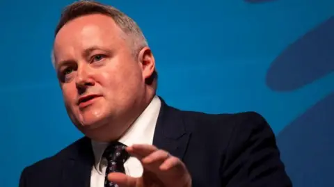 Getty Images Darren Millar giving a speech at a conference with a blue background behind him.