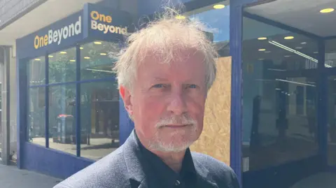 Grace Wood/BBC A man with short white hair and beard stands in front of a boarded up shop