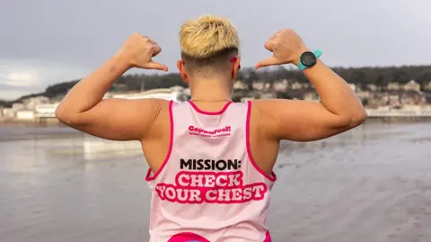 Hana Laurie Sam Smith standing on the beach, pointing at the back of her pink running vest with Coppafeel branding on it, which urges others to check their chest for signs of cancer. 