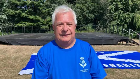 Mark Pengelly, wearing a blue T-shirt, with a flag of Scotland behind him. An empty stage is also set up behind him, with trees and bushes in the distance.
