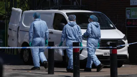 PA Media Three people wearing forensic protective suits, hair coverings and gloves walk beside a white van behind a police cordon. Bollards and tape are visible in the foreground, with a building and fencing in the background.