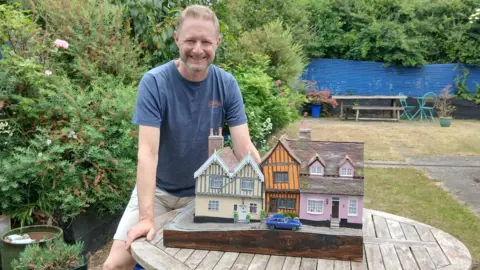 Simon Evans A man poses next to a model of a row of cottages. There are two cottages, one pink and one cream, with a smaller, orange crooked cottage between them. There is a blue car parked outside the orange home. The man has light hair and wears a blue T-shirt and cream shorts. He rests his hands on the table that the model sits on. 