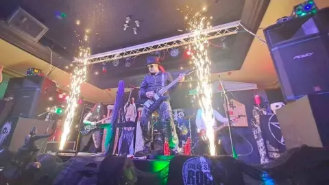 Billibee Creative Studio A man is wearing an all black outfit and a top hat. He is holding a guitar with band members standing behind him as sparklers spray from the stage.