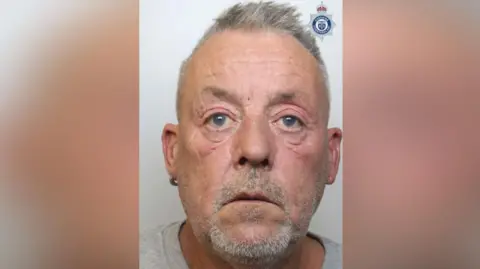 Cheshire Constabulary Gavin Shaw has short grey hair and a short grey beard. He is wearing a grey top and a small hoop earring in his right ear.