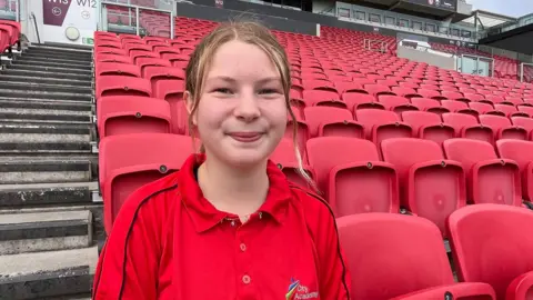 Talia, who has blonde hair pulled back in a ponytail, smiles at the camera. She wears a red City Academy polo shirt, which matches the bank of red seating behind her.