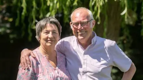 Thames Valley Police Madge and Ed, standing outside, smiling and looking at the camera. Madge is wearing a floral top, with short hair and Ed, has his arm around Madge, wearing a pink shirt, they are standing in front of a tree.
