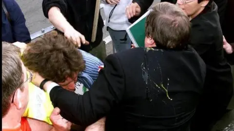 Two men being held back by numerous different hands. Both of the men are grappling for each other. The bystanders have worried expressions on their faces. Egg dripping down the back of one of the man's black suit jacket. 