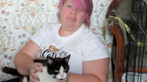 A woman with pink hair wearing a cat T-shirt who is smiling and holding a black and white cat. In the background can be seen a floral sofa, a chair and a cat crate.
