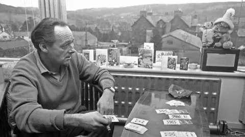 Mirrorpix via Getty Images Donald Campbell plays cards as he holds a cigar in his right hand. He is sitting next to a small wooden table and houses and hills can be seen through the window behind him. One of his mascots, a teddy bear named Mr Whoppit, is placed on the window sill.