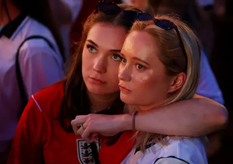 Reuters Two England fans look dejected after the match. One woman in a red England shirt with sunglasses pinned on top of her head has her arm wrapped around another woman in a white top.