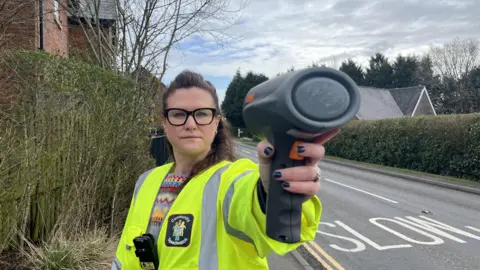 Sarah Jones who has brown hair wearing a high vis with body camera and a radar gun to measure speed standing next to a road with a slow sign painted on on the outskirts of the village