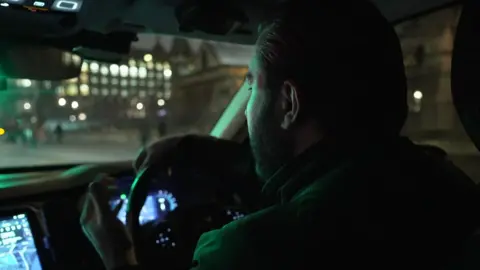 PC Matt Murrant is driving a police car through the West End, it is dark outside and he is turned away from the camera, watching the road 
