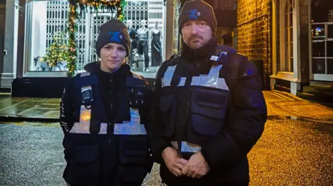 North Yorkshire Council Night marshals stand by a road in Harrogate. They wear black vests with fluorescent silver stripes, and grey beanie hats.