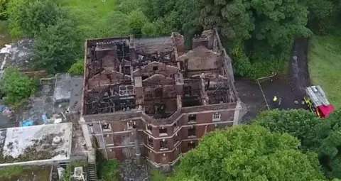 Aerial view of Daresbury Hall after fire ravaged the historic Georgian manor. It has no roof and many of the windows are shot. A fire engine is seen parked to the right of the building.