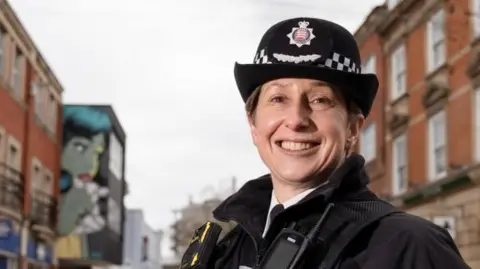 Rachel Nolan smiles at the camera. It is a head-and-shoulders shot. She is wearing her uniform and police hat and is standing in a high street. The street is out of focus.