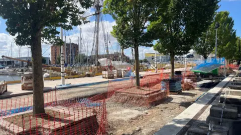A construction site near a waterfront, with red safety netting, stacks of bricks, and wooden pallets. Trees and docked boats are visible in the background, along with buildings and cranes.