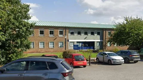 Bishop Auckland Police Station. The picture has been taken from the car park, where several cars are parked. Behind them is the police station, which has a green corregated rood and brown brick walls. The entrance is blue.