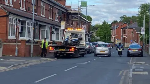 BBC Blue delivery moped on the back of a lorry at the side of Cromwell Grove, which is lined by red brick terraced houses.