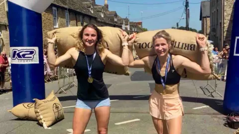 Two young women in shorts and sports tops pose with woolsacks on their shoulders after completing the Tetbury Woolsack Races. 