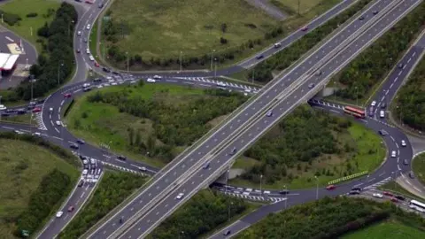 A mocked-up image of two dual carriageways running over a large roundabout with trees and vegetation in the middle. A building's roof can be seen to the left of the image.