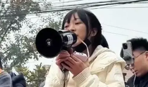 Jimu News A girl wearing a white top clutching a mobile phone users a black loudspeaker. A tree and a man with a phone seemingly attached to his head are in the background 