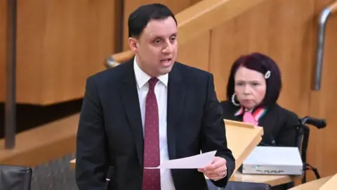 Anas Sarwar, who has short black hair, speaks in the Scottish Parliament chamber. He is holding a piece of paper in his left hand and wearing a dark suit, white shirt and red tie. 