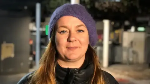 Carly Heath looks straight on. She has green eyes and light brown hair. She wears a black jacket and a purple woolly hat. It is night-time and the light from the street lights shines on her eyes as she gives a half-smile.