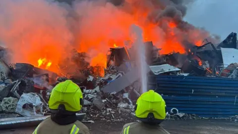 Two firefighters in brown gear with yellow reflective stripes and yellow helmets are standing in front of a large pile of metal waste. It is engulfed in orange flames and has grey and black smoke billowing from it. One has a hose of water and is spraying it at the fire