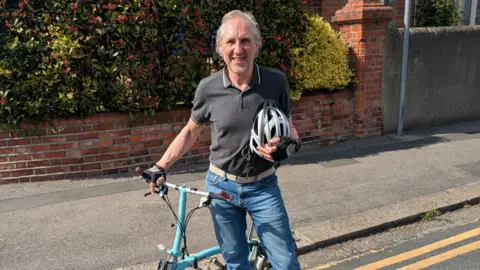 Tony Carr A man smiling into the camera while stood next to a bike and holding a helmet.